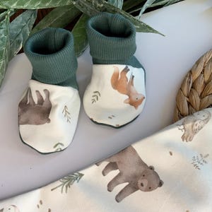 May include: A pair of green and white baby booties with woodland animal designs. One bootie features a brown bear and the other a fox.