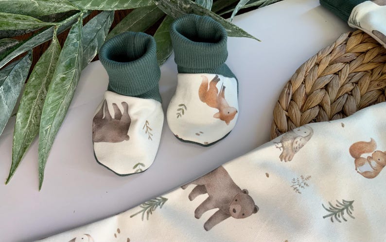 May include: A pair of green and white baby booties with woodland animal designs. One bootie features a brown bear and the other a fox.