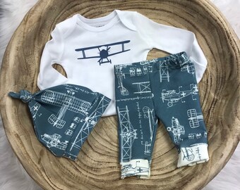 May include: A baby outfit set with a white long-sleeved onesie featuring a blue airplane graphic, blue pants with a white airplane print, and a blue hat with a white airplane print.