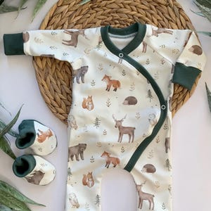 May include: A white baby onesie with a green trim and a woodland animal print. The onesie has a wrap-around design and is shown with a pair of green baby booties.