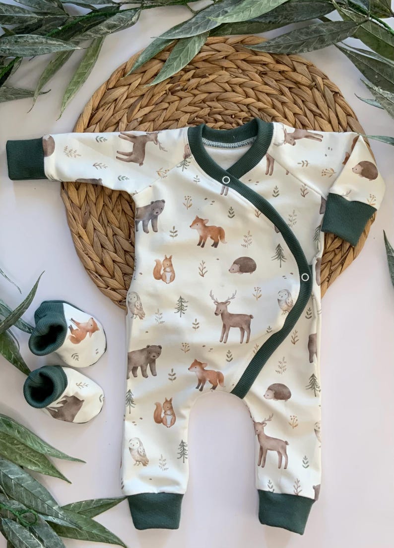 May include: A white baby onesie with a green trim and a woodland animal print. The onesie has a wrap-around design and is shown with a pair of green baby booties.