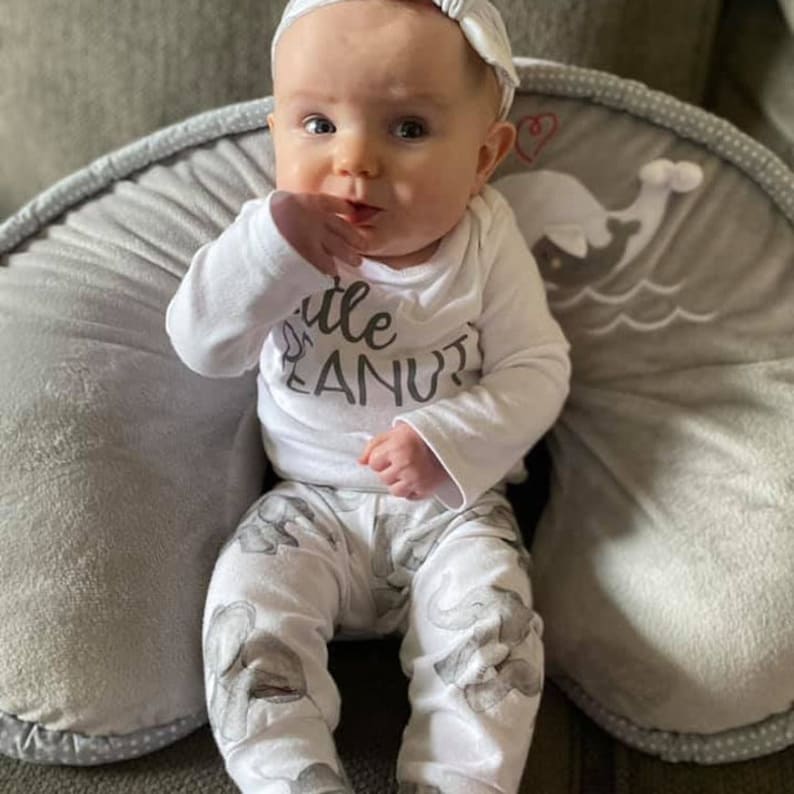 May include: A baby wearing a white onesie with the text "Little Peanut" and white pants with a gray elephant print sits on a gray and white pillow.