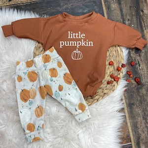 Könnte beinhalten: Ein braunes Langarmshirt mit dem Text "little pumpkin" und einer kleinen Kürbisgrafik. Eine weiße Hose mit einem Kürbisdruck.