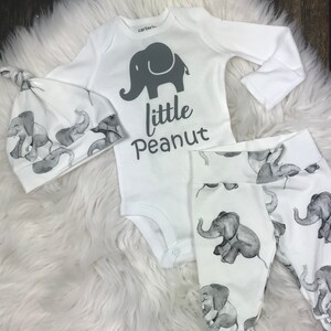 K&ouml;nnte beinhalten: Ein wei&szlig;es Baby-Outfit-Set mit Elefanten-Druck. Das Set beinhaltet einen lang&auml;rmeligen Strampler, eine M&uuml;tze und Leggings. Der Strampler hat ein graues Elefanten-Motiv und den Text "little peanut".