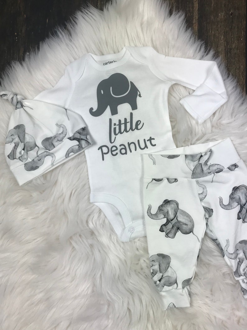 K&ouml;nnte beinhalten: Ein wei&szlig;es Baby-Outfit-Set mit Elefanten-Druck. Das Set beinhaltet einen lang&auml;rmeligen Strampler, eine M&uuml;tze und Leggings. Der Strampler hat ein graues Elefanten-Motiv und den Text "little peanut".