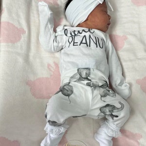 May include: A white baby onesie with the text "little peanut" printed on the front. The onesie is paired with white pants featuring a gray elephant print. The baby is wearing a white headband.