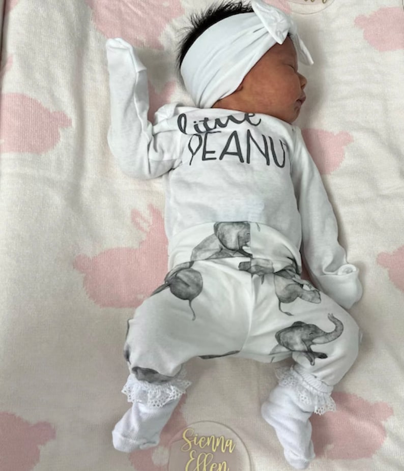 May include: A white baby onesie with the text "little peanut" printed on the front. The onesie is paired with white pants featuring a gray elephant print. The baby is wearing a white headband.