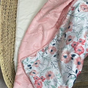 May include: A pink and white baby blanket with a floral pattern. One side is a soft, plush pink fabric, and the other side is a white fabric with pink flowers and green leaves.