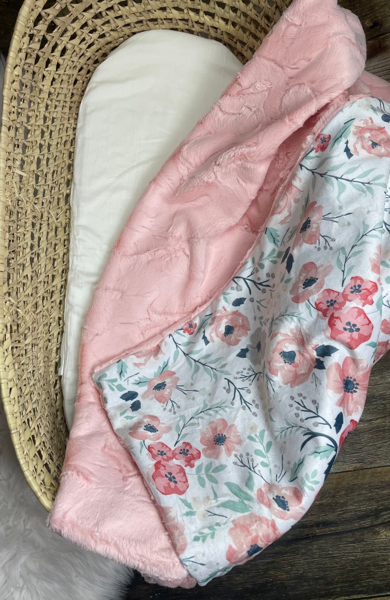 May include: A pink and white baby blanket with a floral pattern. One side is a soft, plush pink fabric, and the other side is a white fabric with pink flowers and green leaves.