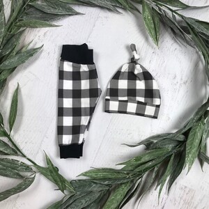 May include: Black and white checked baby trousers and hat set. The trousers have a black elastic waistband and the hat has a knotted top.