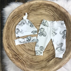 K&ouml;nnte beinhalten: Ein wei&szlig;es Baby-Outfit-Set mit Elefanten-Druck. Das Set beinhaltet eine M&uuml;tze, Leggings und ein Stirnband.
