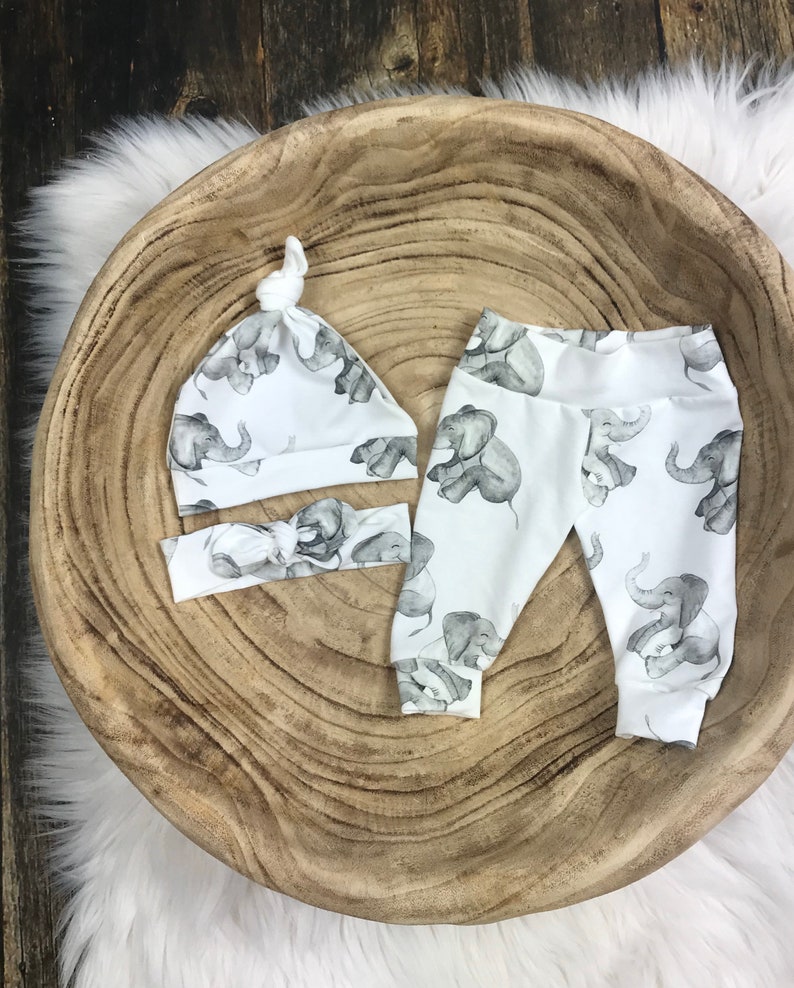 K&ouml;nnte beinhalten: Ein wei&szlig;es Baby-Outfit-Set mit Elefanten-Druck. Das Set beinhaltet eine M&uuml;tze, Leggings und ein Stirnband.