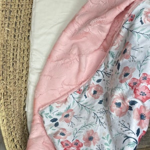 May include: A pink and white baby blanket with a floral pattern. The blanket is made of soft, plush fabric and is perfect for cuddling.