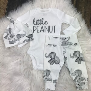 May include: A white baby outfit with a long-sleeved bodysuit, pants, and a hat. The bodysuit has the text "little peanut" printed on it. The pants and hat have a gray elephant print.