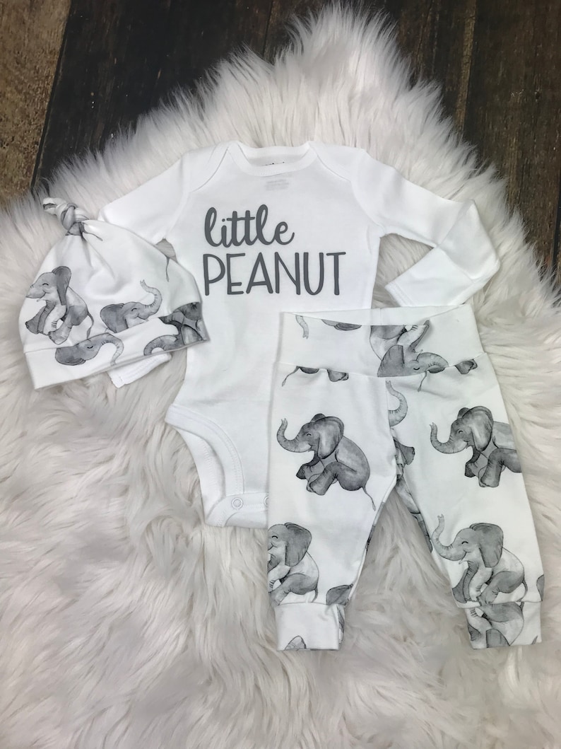 May include: A white baby outfit with a long-sleeved bodysuit, pants, and a hat. The bodysuit has the text "little peanut" printed on it. The pants and hat have a gray elephant print.