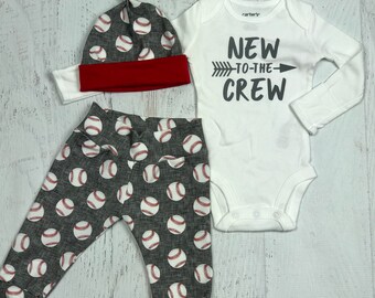 May include: A baby outfit set with a white long-sleeve bodysuit with the text "NEW TO THE CREW" and an arrow, gray and white baseball print pants, and a gray and white baseball print hat with a red brim.