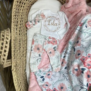 May include: A baby outfit set with a white long-sleeved onesie with the name "Isla" embroidered on it, pink floral leggings, a pink headband, and a pink and white floral blanket.