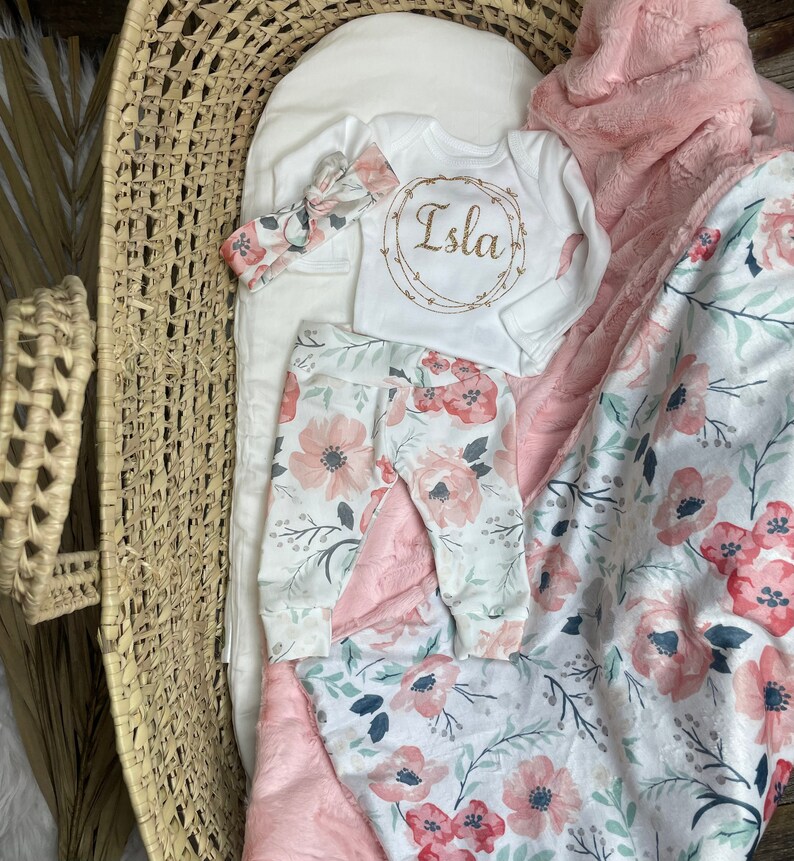 May include: A baby outfit set with a white long-sleeved onesie with the name "Isla" embroidered on it, pink floral leggings, a pink headband, and a pink and white floral blanket.