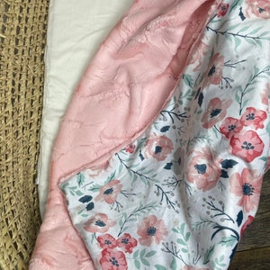 May include: A pink and white baby blanket with a floral pattern. One side of the blanket is soft and fuzzy, while the other side is made of a smooth fabric.