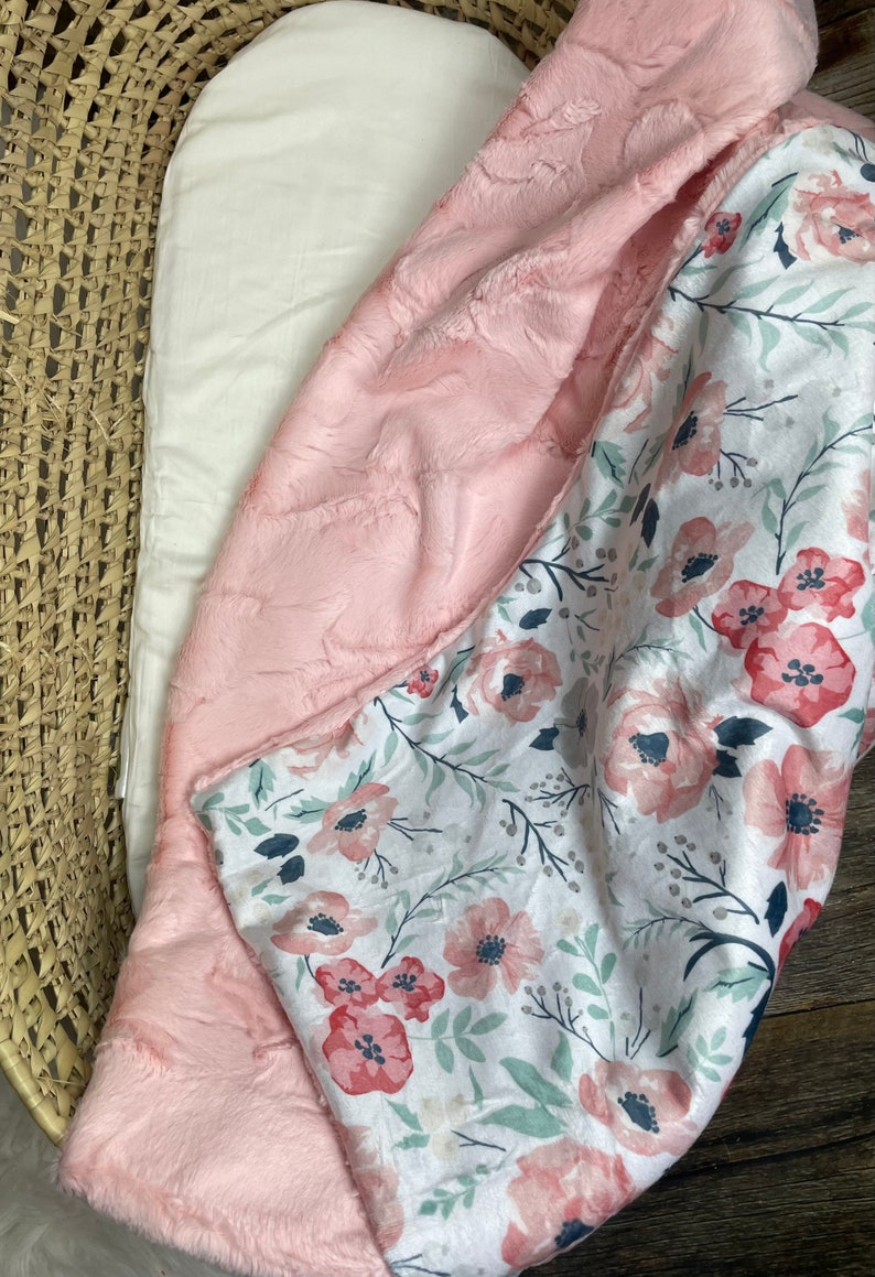 May include: A pink and white baby blanket with a floral pattern. One side of the blanket is soft and fuzzy, while the other side is made of a smooth fabric.