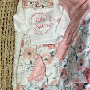 May include: A white baby onesie with the text "Little Sister" in a red circle with vines. The onesie is paired with floral print pants and a pink floral print blanket.