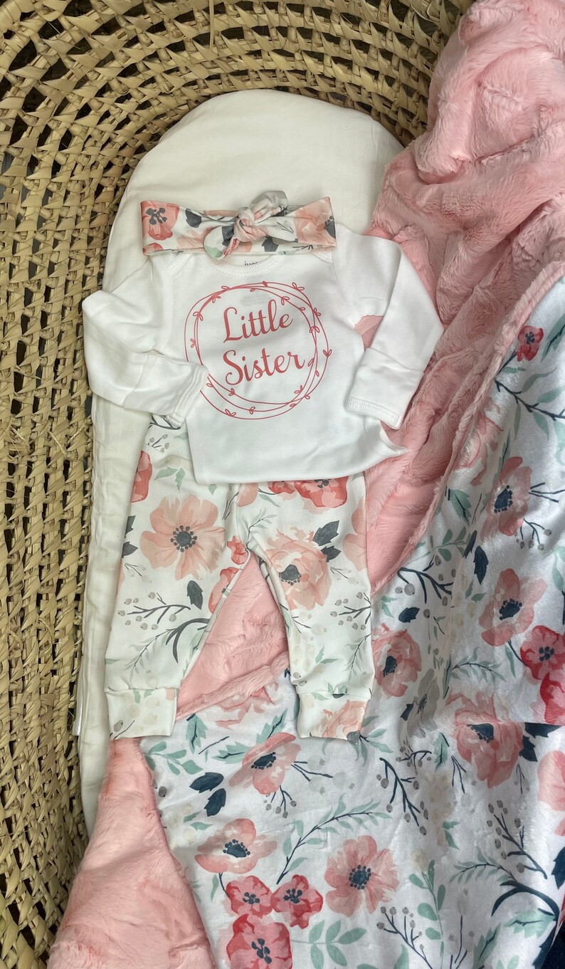 May include: A white baby onesie with the text "Little Sister" in a red circle with vines. The onesie is paired with floral print pants and a pink floral print blanket.