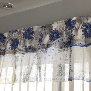 May include: A white valance with a blue floral pattern. The valance is made of two layers of fabric, with the top layer featuring a larger floral design and the bottom layer featuring a smaller floral design. The valance is gathered at the top and has a ruffled edge.