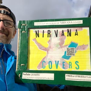 May include: A green cardboard sign displaying the words "NIRVANA COVERS" in bold, colourful letters. The sign also features the text "CHECK OUT MY ALBUM OF NIRVANA COVERS" and social media details. A QR code is visible.