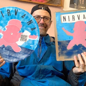 May include: Two Nirvana-themed artworks are held up. One is a painted vinyl record with a pink figure and the band's name in blue. The other is a cardboard piece with the same design. Both feature a QR code.