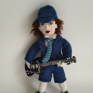 May include: A knitted doll wearing a blue suit, a blue cap, and a blue and white tie. The doll is holding a black electric guitar.