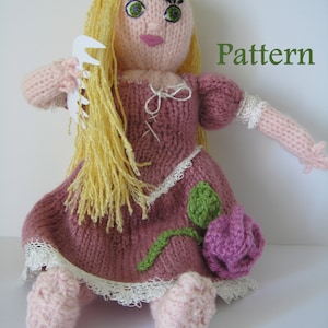May include: A knitted doll with long blonde hair, wearing a pink dress with a white lace trim and a green leaf and purple rose detail. The doll is holding a white comb in her hand. The text "Pattern" is written in green letters at the top of the image.