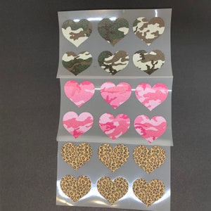 May include: A sheet of heart-shaped stickers in various patterns. The patterns include camouflage in green and brown, pink camouflage, and leopard print. The stickers are arranged in rows and columns on a clear backing.