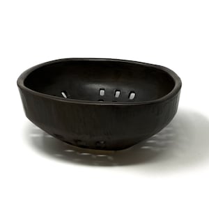May include: A black ceramic bowl with a unique design featuring a series of holes around the rim. The bowl is perfect for serving fruit, snacks, or other small items.