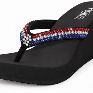 May include: A black wedge sandal featuring a thong-style strap adorned with rows of red, white, and blue rhinestones. The footbed is solid black with the brand name Flojos printed in white text near the heel area of the wedge sole.