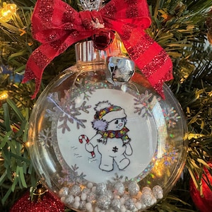May include: A clear glass Christmas ornament with a white background featuring a cartoon snowman wearing a purple and yellow scarf and a blue hat. The snowman is holding a candy cane and is surrounded by silver glitter snowflakes. The ornament is decorated with a red and white striped bow and a silver bell.