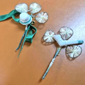May include: Two vintage brooches with a floral design. Each brooch features three cream-colored, circular elements with a dark outline. One has a small white hat and green ribbon, the other has a white element.