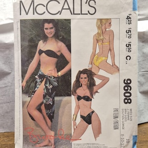 May include: A McCall's 9608 sewing pattern envelope featuring a photograph and illustrations of a two-piece swimsuit. The design includes a strapless bandeau top and a bikini bottom, with an optional sarong wrap featuring a dark floral print.
