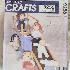 Könnte beinhalten: McCall's Crafts 9256 Schnittmusterumschlag für 41 cm große Stoffpuppen. Das Cover zeigt Illustrationen von fünf Stoffpuppen in verschiedenen Outfits auf einer hölzernen Klettergerüststruktur, mit dem handschriftlichen Namen Sandy auf der Vorderseite.