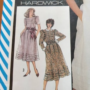 Può includere: Illustrazione di due abiti da un modello di cucito Hardwick. Uno è lilla chiaro con cintura abbinata. L'altro è una stampa floreale con colletto e polsini bianchi. Gli abiti hanno dettagli a pieghe.