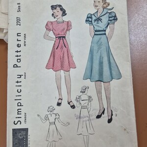 Op de afbeelding: Vintage Simplicity patroon 2707, maat 8, met jurk illustraties. De omslag toont een roze polkadot jurk en een blauwe jurk met pofmouwen. Het patroon bevat naaiaanwijzingen, met vermelding van plaatsen als Parijs, Londen en New York.