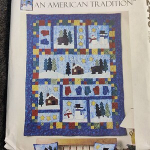 May include: A quilted wall hanging with a winter scene. The quilt features snowmen, bears, trees, houses, and mittens in various squares. The border is blue with a patchwork design. The text "AN AMERICAN TRADITION" is at the top.