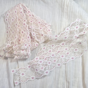 May include: Off-white lace trim with a delicate floral pattern in pink. The lace is folded and laid out on a white fabric background. This decorative trim is suitable for sewing and craft projects.