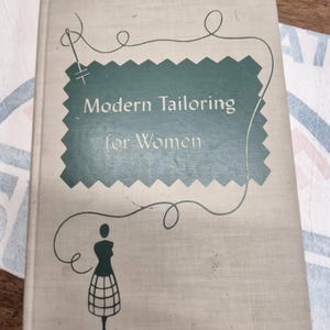 以下が含まれることがあります： 「Modern Tailoring for Women」というタイトルのヴィンテージ本。薄いベージュ色の表紙で、タイトルは濃い緑色のテクスチャのある長方形の中にあります。スタイリッシュなマネキンと針と糸のデザインも表紙にあります。