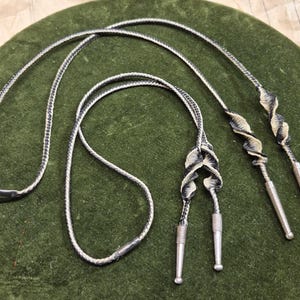 May include: A bolo tie featuring a braided black and white cord with silver-toned metal tips. The cord is accented with two decorative, twisted black and white spiral elements that lead to four silver-toned metal aglets at the ends of the cord.