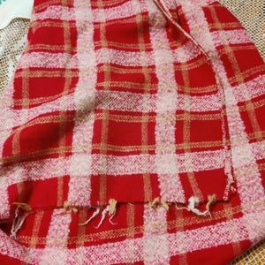 May include: A red and white plaid textile with a fringed edge. The pattern features a grid of red squares and rectangles, with beige and white accents. The fabric appears to be woven.