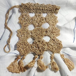 May include: A tan-colored, open-weave drawstring bag with a decorative floral pattern. The bag features a series of tassels along the bottom edge and a woven drawstring. The bag is made of a natural fiber.