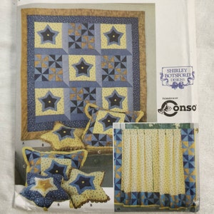 May include: A quilt and star-shaped pillows with a matching curtain pattern. The quilt features large yellow stars on a blue background, with a brown border. The pillows and curtain have a similar design. The image includes the text "Shirley Botsford Designs".