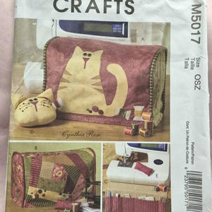 May include: McCall's Crafts sewing pattern featuring a cat design. The pattern includes instructions for a sewing machine cover and other accessories. The main image shows a cat-themed cover in burgundy and tan, with additional design options.