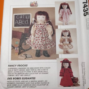 May include: A sewing pattern for Emma doll clothing, showcasing various doll outfits. The main doll is in a strawberry-print dress and black boots. Other outfits include a pink dress, a tan dress, and a red coat with a matching hat. The pattern fits 45cm dolls.