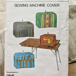 May include: A white paper sewing pattern envelope labelled SIRO SEWING MACHINE COVER. The front displays four illustrations of sewing machine covers labelled A, B, C, and D, featuring various patterns, colours, and storage pockets on a wooden table.